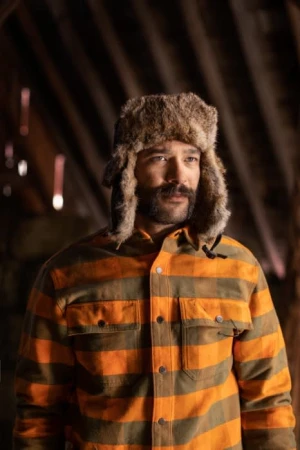 Man in fur hat and striped shirt in rustic interior, looking off-camera. Natural light highlights textures and warm tones.