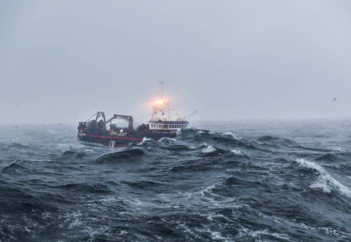 Fishing trawler navigates turbulent dark blue ocean under overcast sky, battling rough seas and weather for a challenging catch.