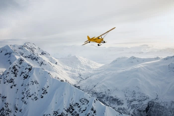 Yellow airplane flying over snow-covered mountains on a partly cloudy day, creating a scenic aerial view of nature's beauty.