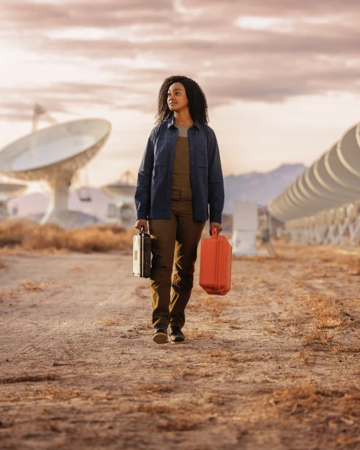 Adult female walking on a dirt road carrying cases, with large satellite dishes visible in the background on a cloudy day.