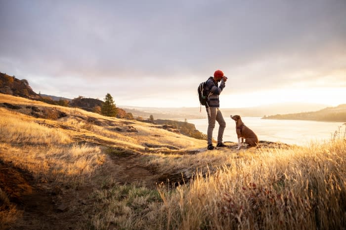 Man and dog sit on a grassy hill overlooking a lake at sunset. The sky is cloudy. Man wears a backpack and a red hat.