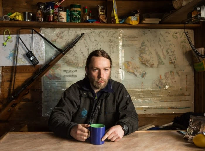 Bearded man in a dark jacket holding a blue mug sits at a table indoors with a rifle and map on the wall behind him.