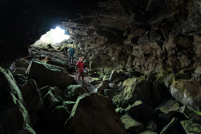Two people explore a moss-covered rocky cave, illuminated by natural light from the cave's entrance, creating a sense of adventure.