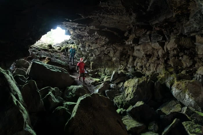 Two people explore a moss-covered rocky cave, illuminated by natural light from the cave's entrance, creating a sense of adventure.
