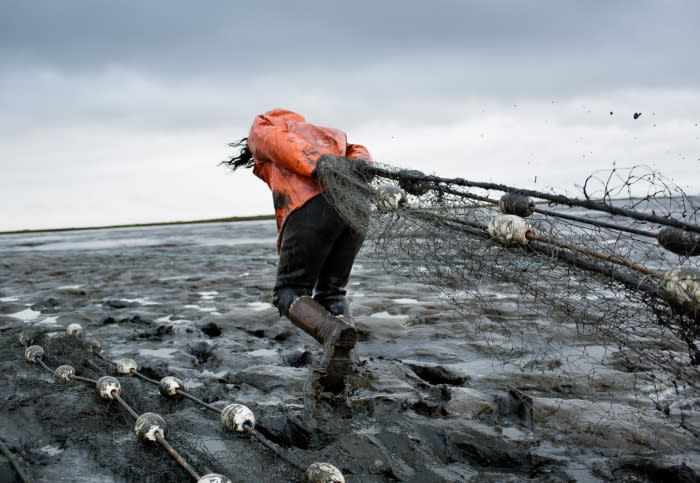 Person in orange jacket pulling fishing net on muddy shore against an overcast sky, wearing boots, capturing the essence of hard work.