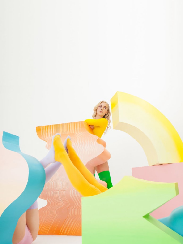 Blonde woman poses playfully among colorful geometric shapes in a studio setting, wearing yellow and green legwear, and a long-sleeved shirt.