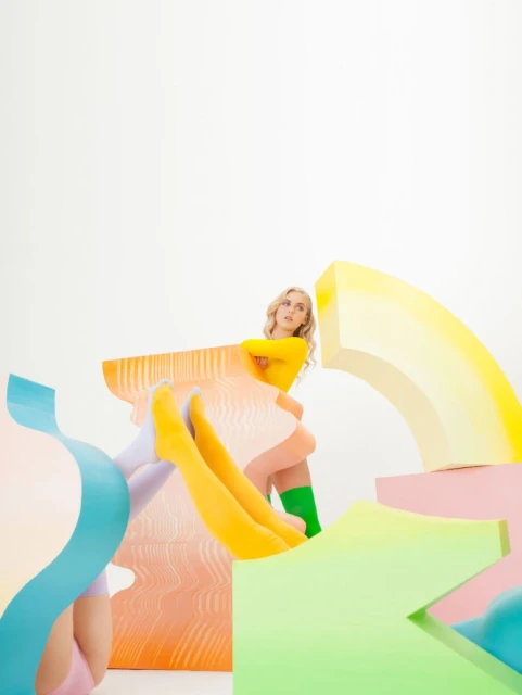 Blonde woman poses playfully among colorful geometric shapes in a studio setting, wearing yellow and green legwear, and a long-sleeved shirt.