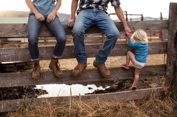 Adults and child on a wooden fence in a rural setting with dry grass and a field in the background on a sunny day.