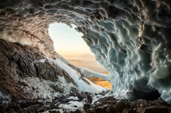 Scenic view inside an ice cave featuring rugged rock, smooth ice formations, and a distant mountain range at sunrise.