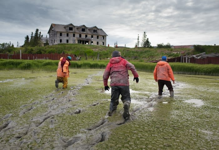 Three people walk through a muddy field toward a large, dilapidated building under a cloudy sky, leaving footprints behind them.