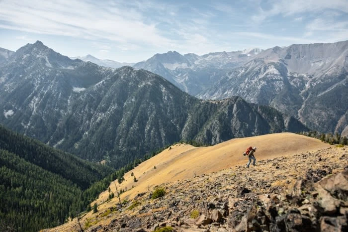 Hiker with backpack walking on a rocky trail near a mountain ridge, with mountain peaks and forest visible under a blue sky.