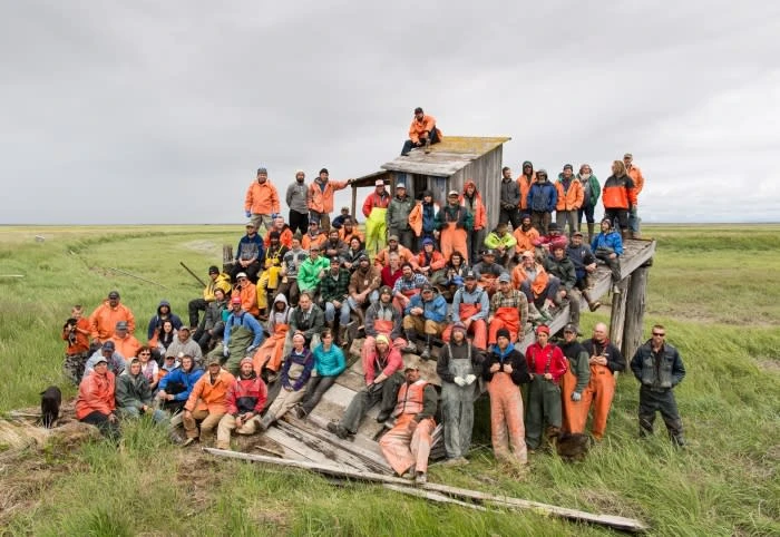 Large group of adults and children posing on and around weathered wooden structure in grassy field under an overcast sky.