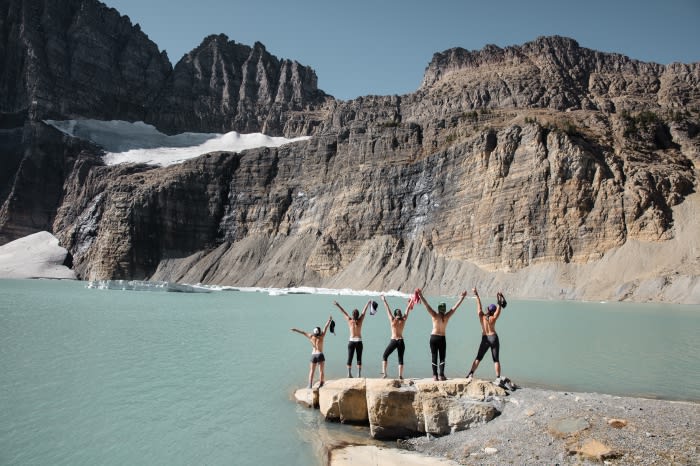 Group of five people stands on a rocky shore with arms raised against a backdrop of a lake and mountainous landscape under a clear sky.