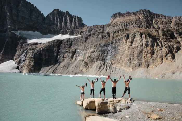 Group of five people stands on a rocky shore with arms raised against a backdrop of a lake and mountainous landscape under a clear sky.