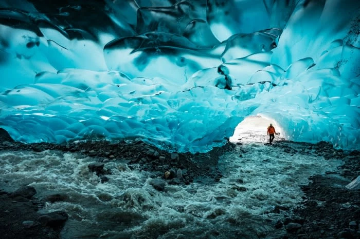 Adult in orange jacket walking through a turquoise-colored ice cave with glacial river flowing along the rocky floor, creating a frozen landscape.