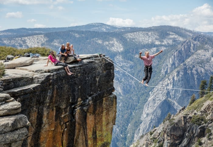 A group watches a man in a pink shirt slacklining across a mountain chasm, featuring rock formations, trees, and blue skies.
