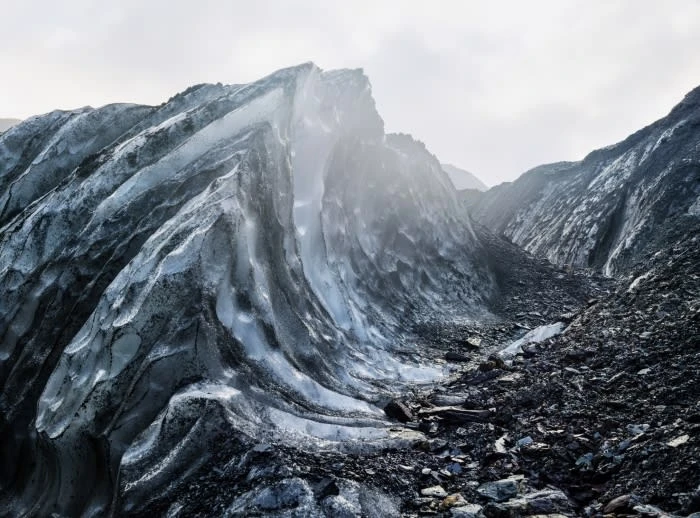 Detailed view of a glacier with blue ice formations and dark rock terrain on a cloudy day, showcasing nature's textures and patterns.