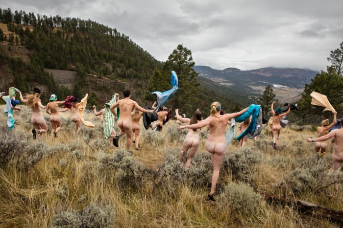 Nude group of women and men running with colorful fabrics in a meadow, mountains and overcast sky in background, captured with wide shot.