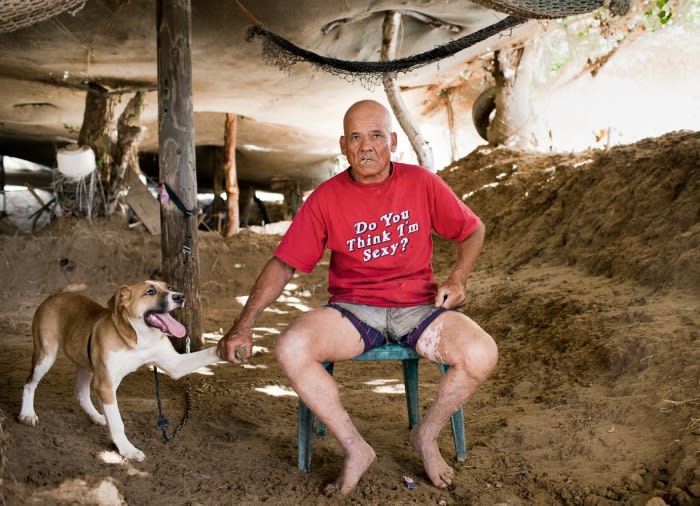 Bald adult man in a red t-shirt sitting on a chair with his dog outside a rustic building with dirt ground.