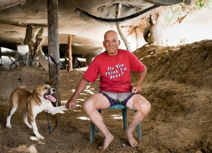 Bald adult man in a red t-shirt sitting on a chair with his dog outside a rustic building with dirt ground.