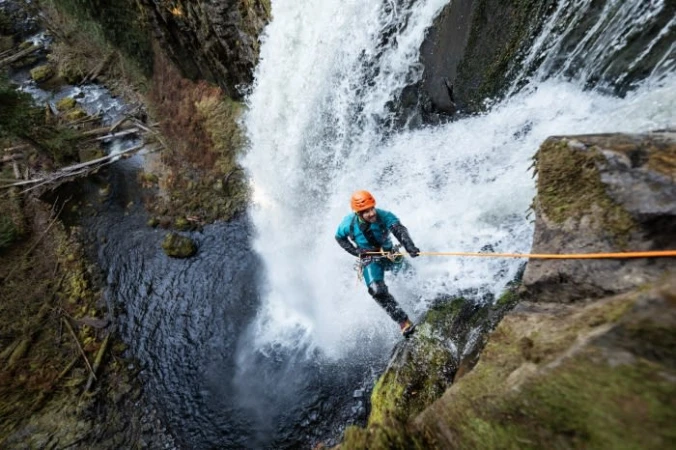A person rappelling down a rock face near a waterfall, wearing protective gear and an orange helmet, surrounded by lush greenery.