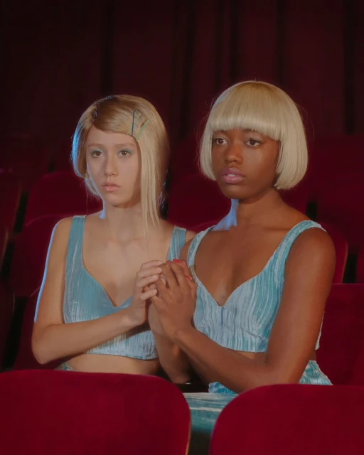 Two women in matching outfits sitting in red theater seats. They have blonde bob haircuts and are holding hands in a medium shot.