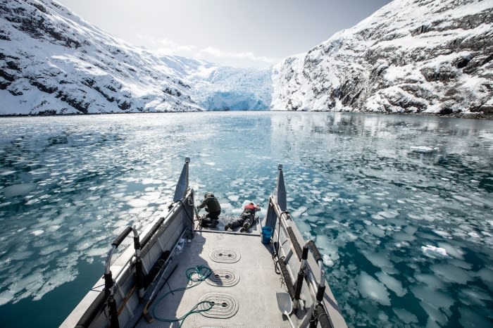 Two people on a boat navigating icy waters, backed by snow-capped mountains and a glacier under a bright sky, landscape orientation.