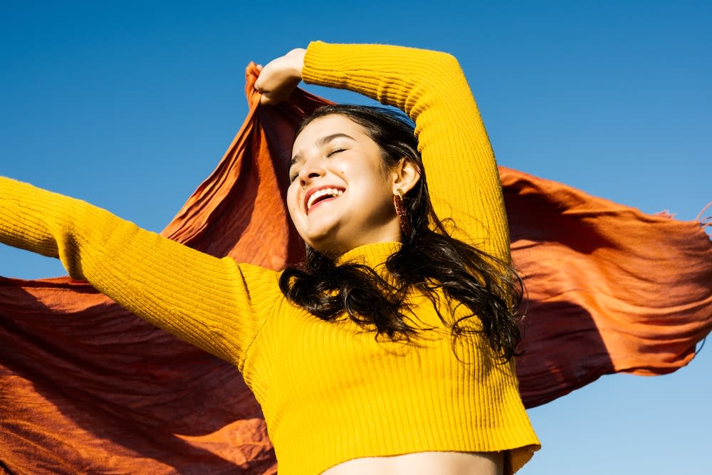 Smiling woman in yellow sweater and orange scarf, arms raised against a bright blue sky. Sunlight illuminates her joyous expression.