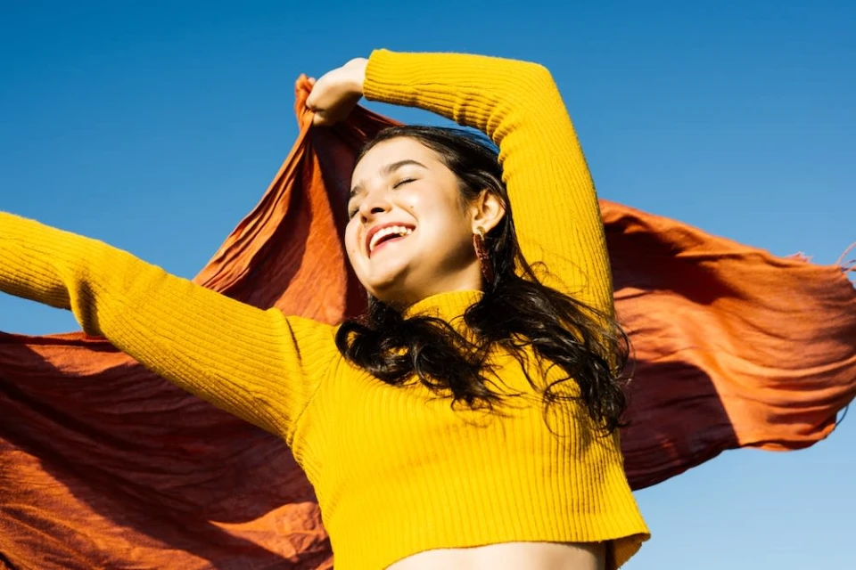 Smiling woman in yellow sweater and orange scarf, arms raised against a bright blue sky. Sunlight illuminates her joyous expression.