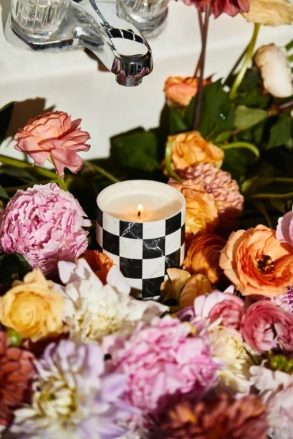Close-up still life of a burning candle surrounded by vibrant pink, orange, and white blossoms next to a silver faucet.