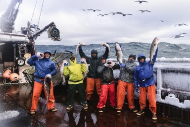 Commercial fishermen in waterproof gear holding up large fish on a frosty deck at sea, photographed by Corey Arnold