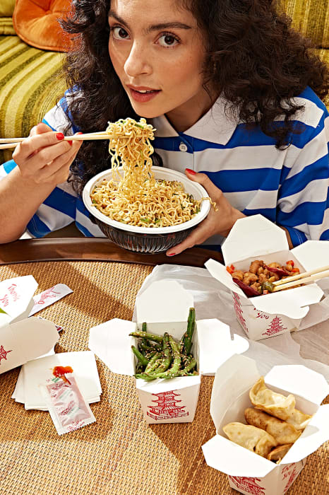 Young woman with dark curly hair enjoys takeout noodles at a table laden with Chinese food containers in a cozy home setting.