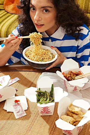 Young woman with dark curly hair enjoys takeout noodles at a table laden with Chinese food containers in a cozy home setting.
