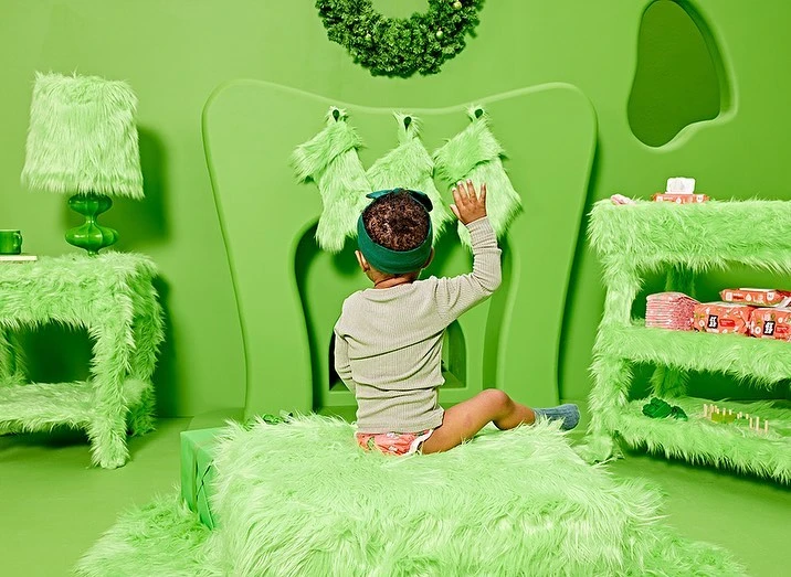 Toddler reaches for Christmas stockings in a whimsical, monochromatic green setting with furry furniture, a rug, and a decorative wreath.
