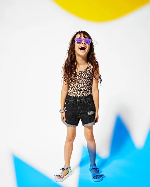 Full-length shot of a young girl smiling with leopard print top, denim shorts, and sunglasses, standing against a vibrant studio backdrop.