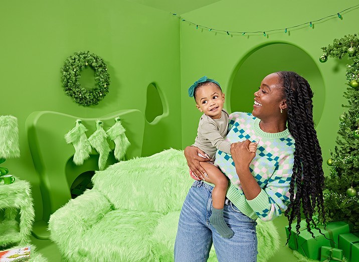 An African-American mother holds her baby girl in a bright green Christmas-themed setting with a tree and whimsical furniture.