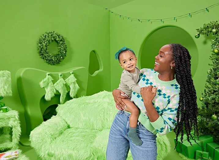 An African-American mother holds her baby girl in a bright green Christmas-themed setting with a tree and whimsical furniture.