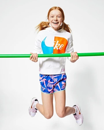 Young red-haired girl jumping while holding a bright green bar, wearing a white hoodie and blue patterned shorts against white background