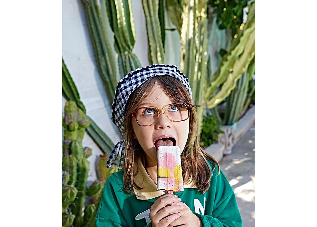 A young child enjoys a multi-colored popsicle outdoors, wearing glasses and a checkered headscarf, with a background of tall cacti.