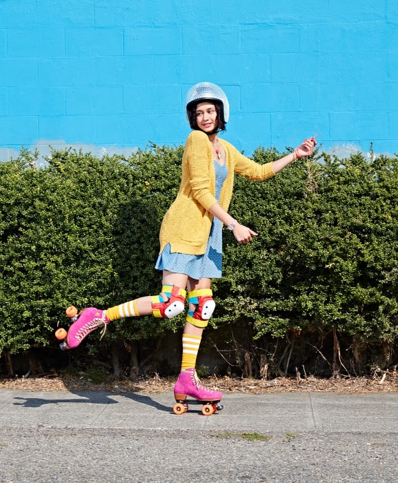 Woman on roller skates wearing protective gear and a helmet in front of a blue wall and green hedge on a sunny day outdoors.