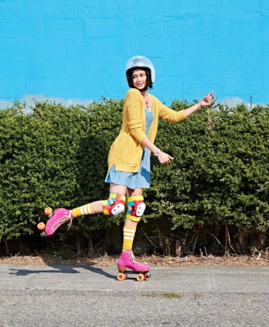 Woman on roller skates wearing protective gear and a helmet in front of a blue wall and green hedge on a sunny day outdoors.