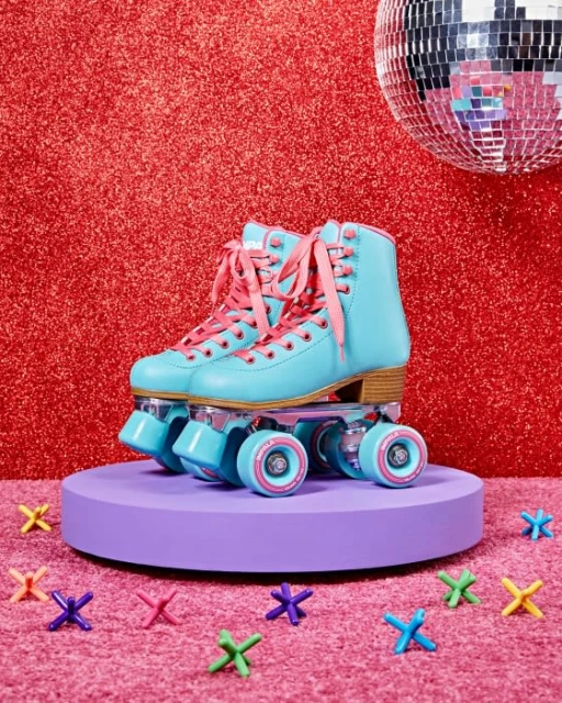A pair of light-blue roller skates with pink laces sit on a purple platform against a red glitter backdrop, surrounded by colorful jacks.