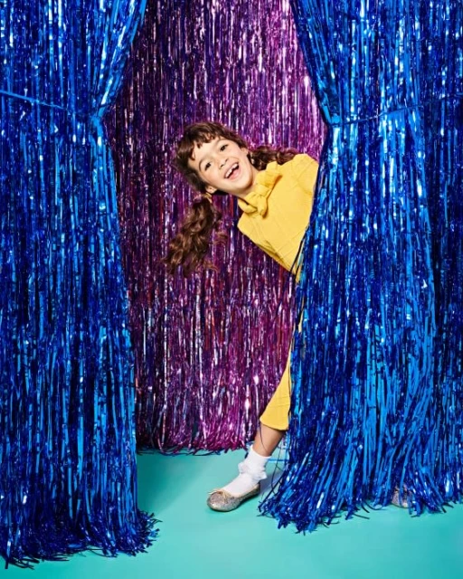 A happy girl with pigtails peeks out from behind blue and purple tinsel curtains against a vibrant turquoise background, smiling broadly.