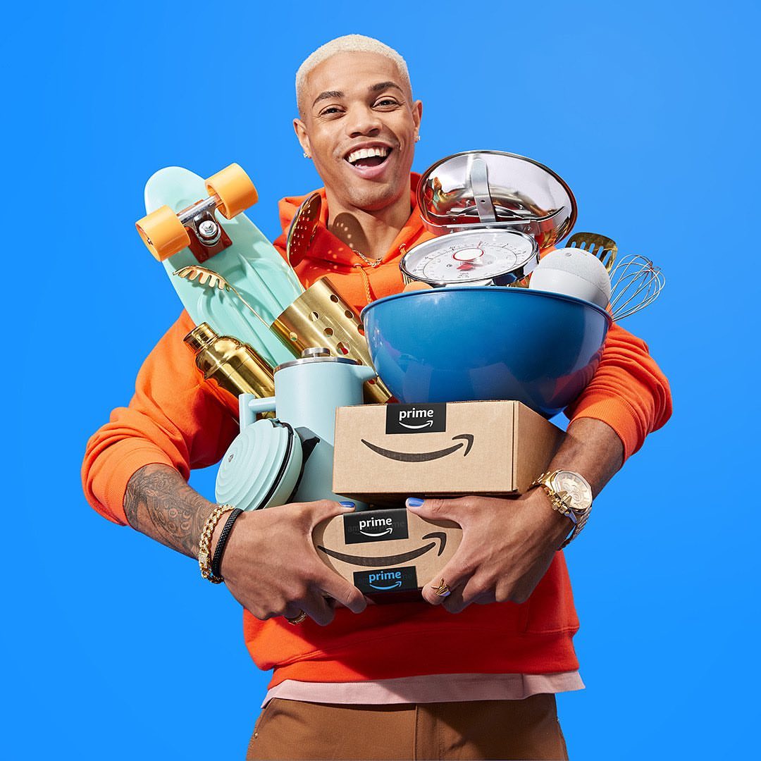 Smiling man with a skateboard and cooking utensils holding Amazon Prime boxes in a studio with a blue background.