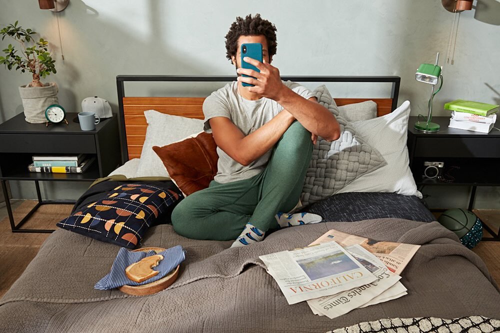 Adult male sits cross-legged on a bed taking a photo of himself with his smartphone in a brightly lit, casual indoor bedroom.