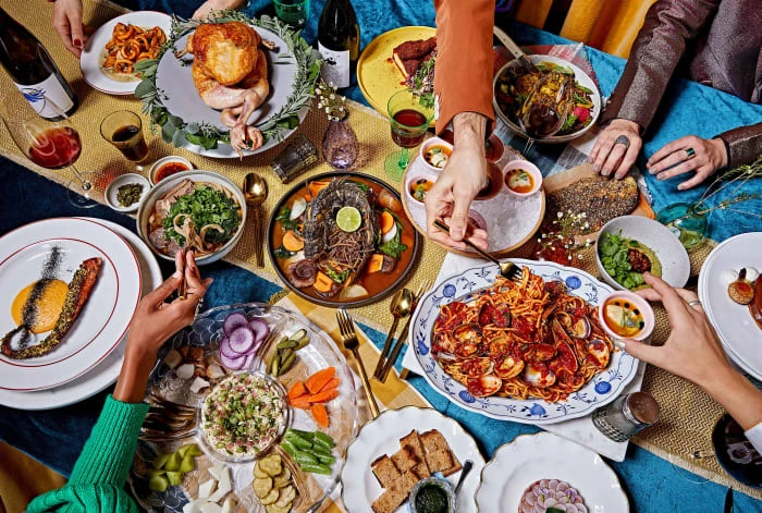 Overhead view of a festive dinner table set with various dishes including roasted chicken, pasta, salad, and appetizers, surrounded by diverse hands.