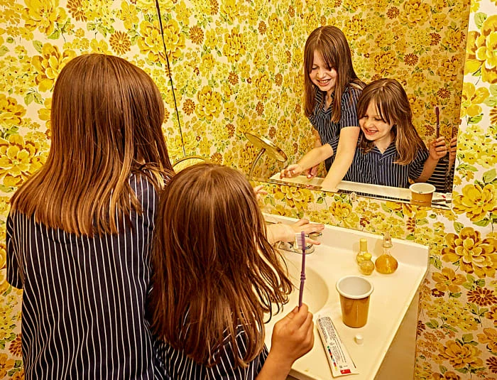 Two young girls brushing their teeth in a bathroom with vintage floral wallpaper, reflected in a mirror with toiletries and a sink.