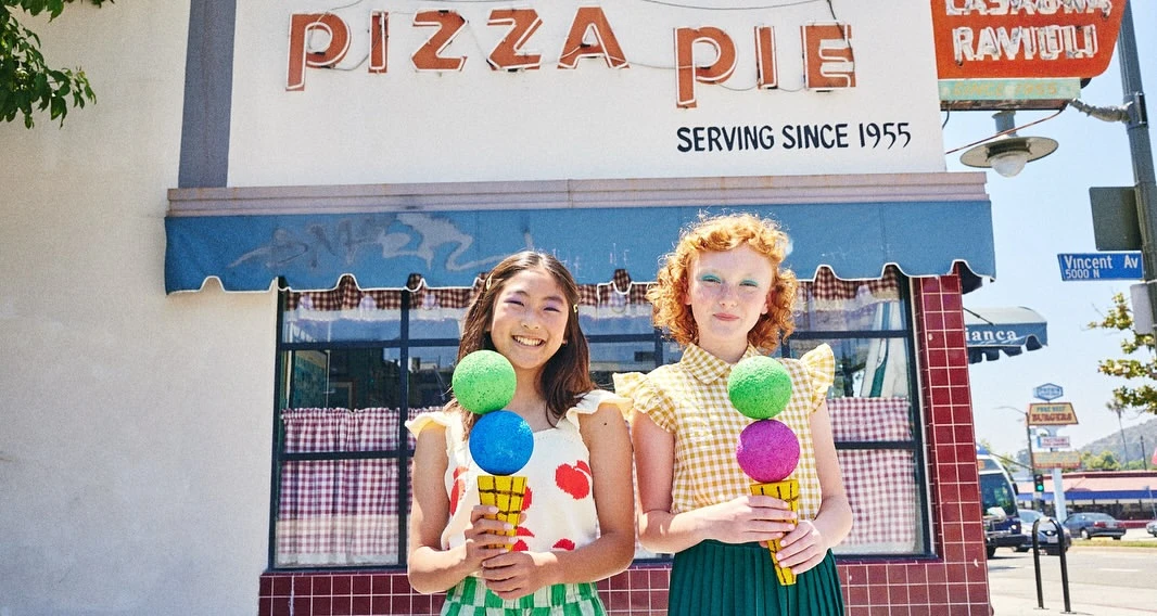 Two young women pose outdoors in front of a Pizza Pie restaurant, holding oversized toy ice cream cones with bright, cheerful expressions.
