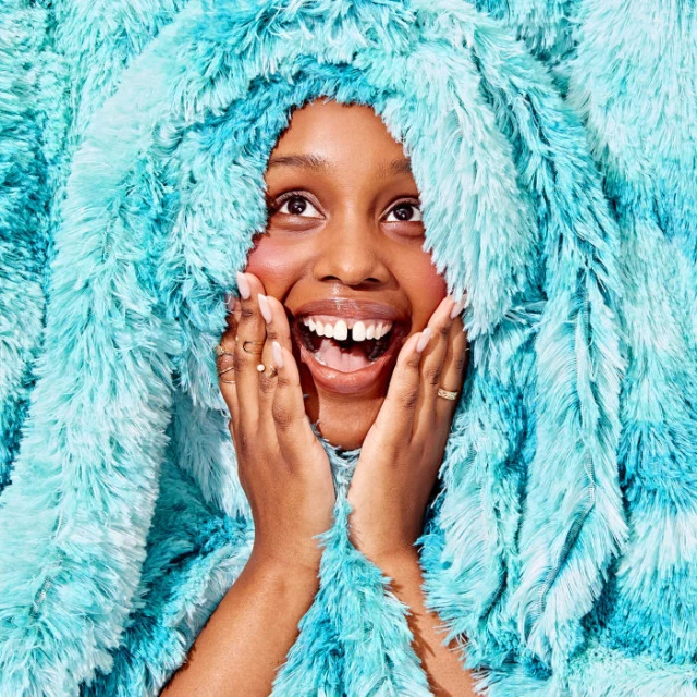 Close-up of a happy Black woman framed by a soft, fluffy blue and white faux-fur hooded blanket, hands on cheeks, looking upwards.