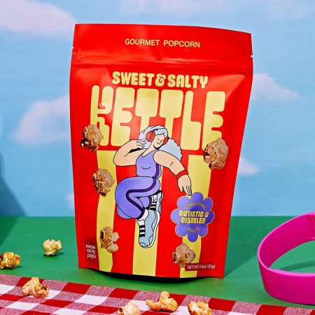 Bright image of a red bag of Sweet & Salty Kettle gourmet popcorn with a whimsical design, set on a checkered tablecloth.